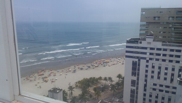 A beach with umbrellas and people, a building with multiple floors, and a view of the ocean. - Beachfront location - Ocean view - Market + Wi-Fi, Cable TV, and Netflix! (Praia Grande)
