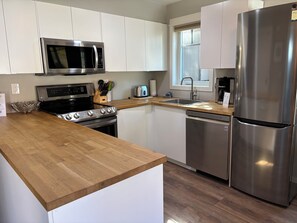 Una cocina moderna con armarios blancos, electrodomésticos de acero inoxidable, encimera de madera y un ventanal que deja entrar luz natural.