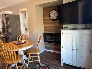 Una cocina moderna con un refrigerador de acero inoxidable, una mesa de comedor de madera, sillas, un reloj y un televisor de pantalla plana montado en la pared.
