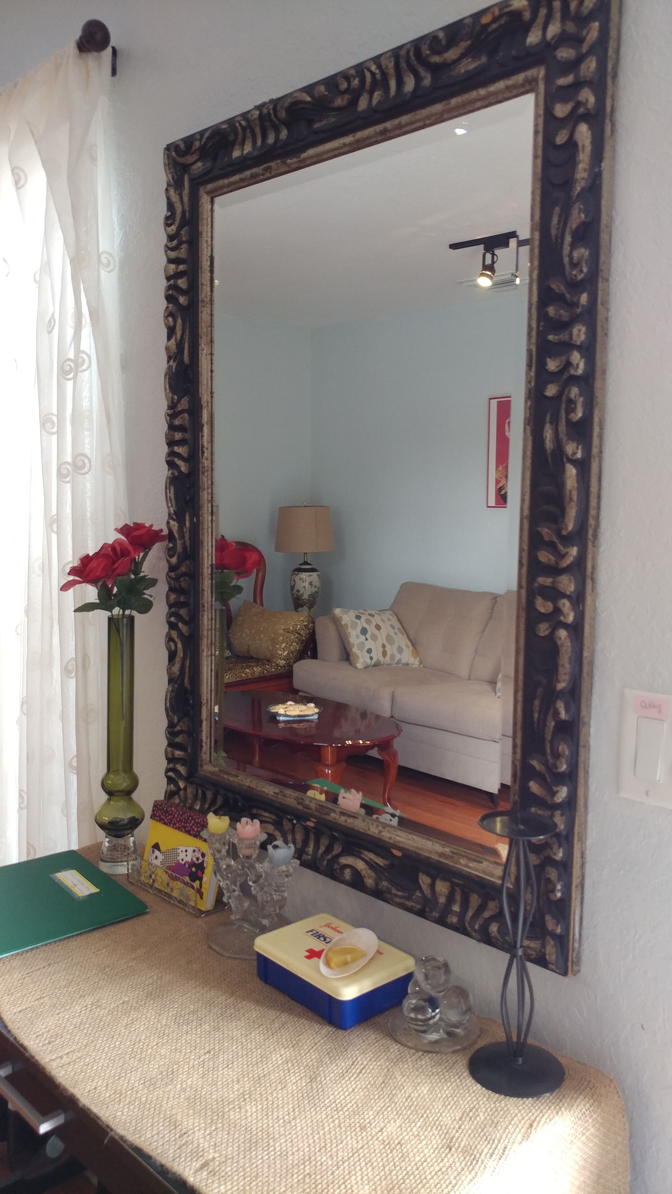 A mirror with a decorative frame, a green vase with red flowers, a lamp, a small table with a box, and a sofa in the background.