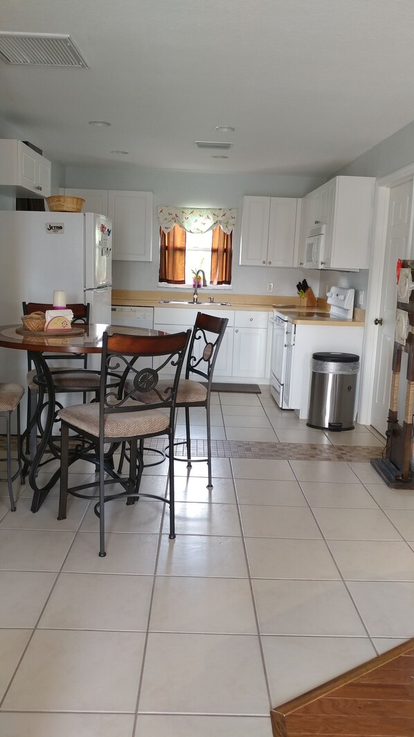 A kitchen with white cabinets, a dining table with chairs, a refrigerator, a sink, and a trash can.