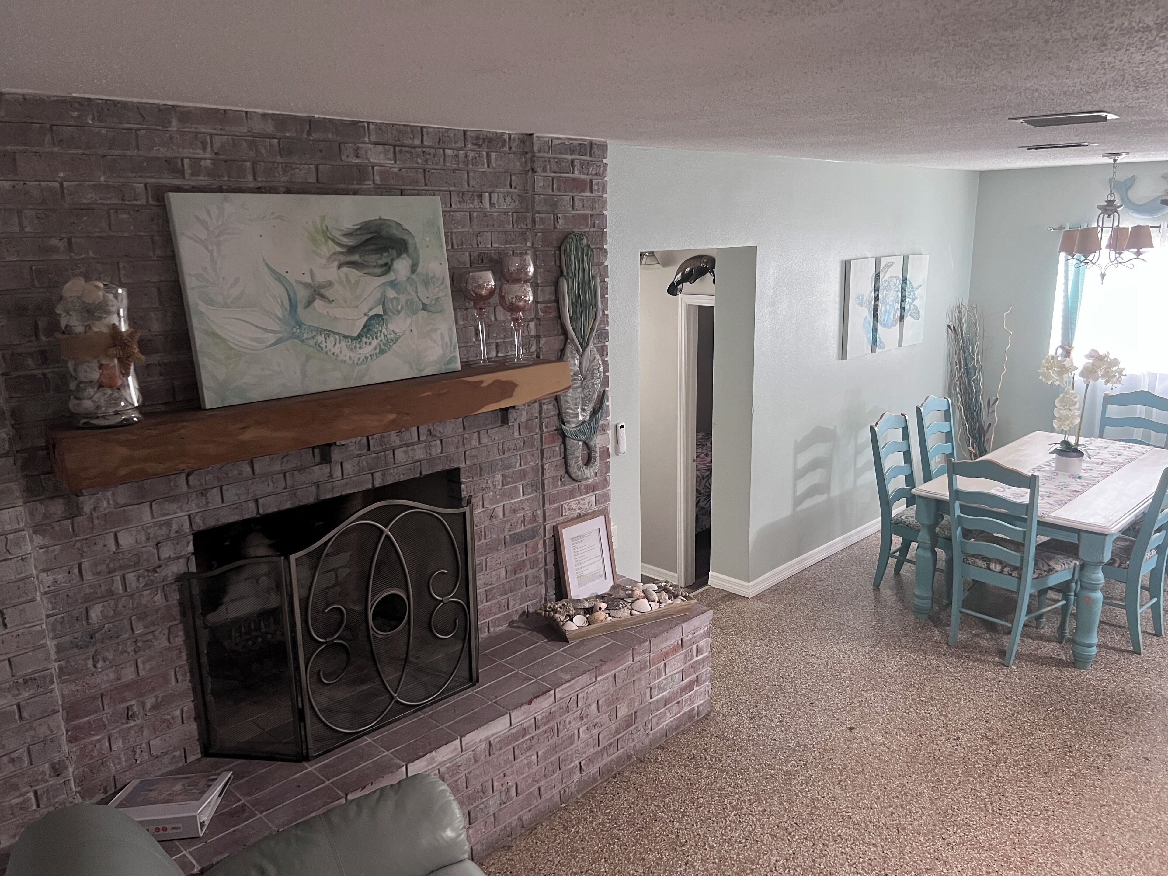 A living room with a brick fireplace, a dining area with blue chairs, and a hallway leading to another room.