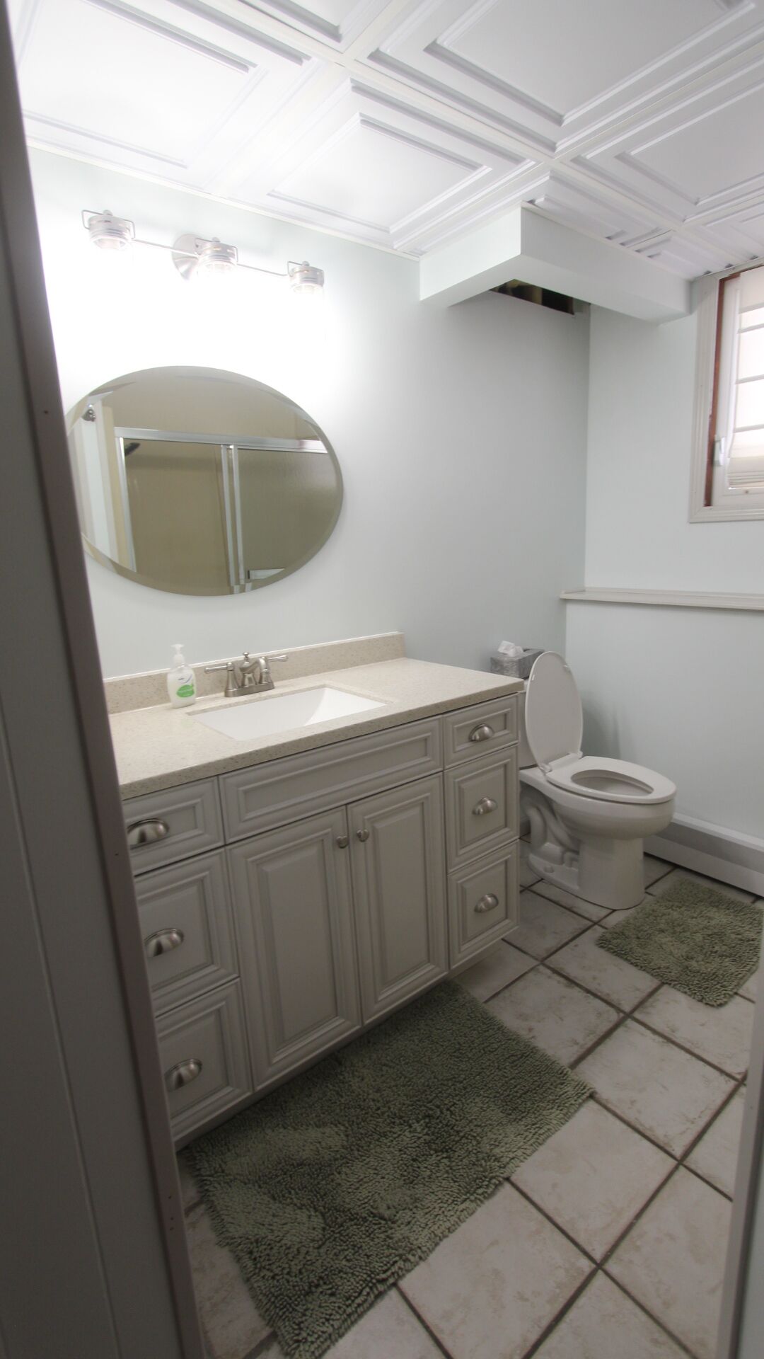 A bathroom with a white vanity, a round mirror, a toilet, and a green rug.