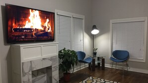 A living room with a fireplace, a TV displaying a fire, two blue chairs, a floor lamp, and a patterned rug. - Historically Modern Delano (Wichita)