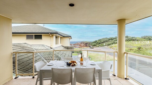 A balcony with a table set for dining, overlooking a coastal landscape with a view of the sea and rocky hills. - Ocean View Anna Bay, 23 Ocean Avenue (Anna Bay)