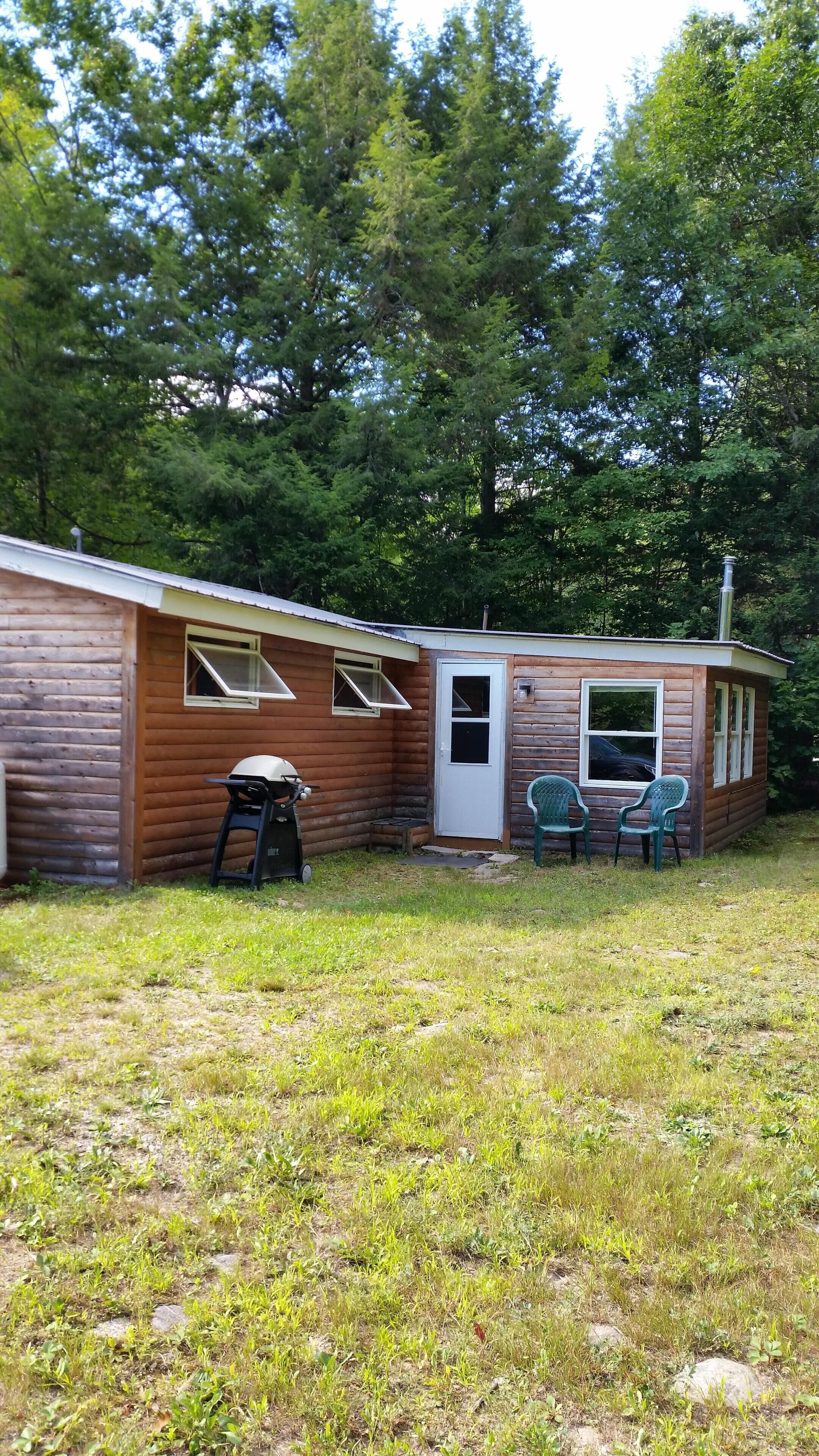 A small wooden cabin with a door, a window, and a small porch area with two chairs.