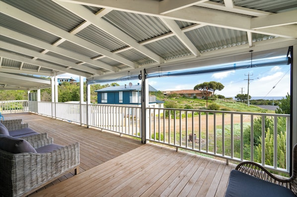 A covered outdoor area with a wooden deck, wicker furniture, and a view of a house and landscape. - Periwinkle By The Beach (Peppermint Grove Beach)
