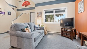 Un salon avec un canapé gris, un téléviseur posé sur un meuble en bois, une fenêtre donnant sur des arbres et un escalier orné d’un éventail rouge.