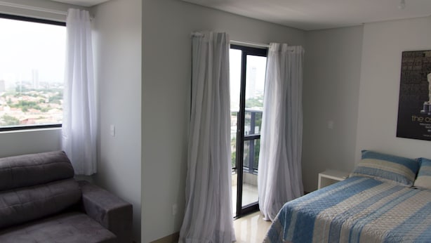Ein modernes Hotelzimmer mit grauem Sofa, einem Bett mit gestreiftem Überwurf und Blick auf die Stadt durch ein großes Fenster mit weißen Vorhängen.