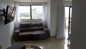 Ein Wohnzimmer mit einer Couch, einem flachbildfernseher an der Wand und einem großen Fenster mit Vorhängen.