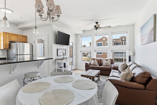 A modern kitchen with a refrigerator, a dining table with placemats, a brown leather sofa, and a ceiling fan.