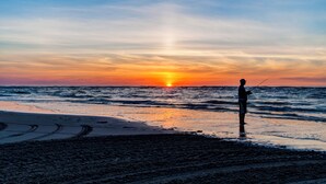 Escena de playa con atardecer, una persona pescando y cielo despejado.