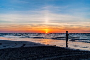 A beach scene with a sunset, a person fishing, and a clear sky.