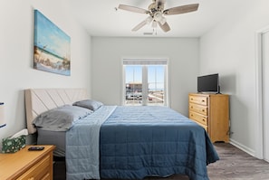 Une chambre à coucher avec un lit, une commode, un téléviseur, un ventilateur de plafond, un tableau et une fenêtre.