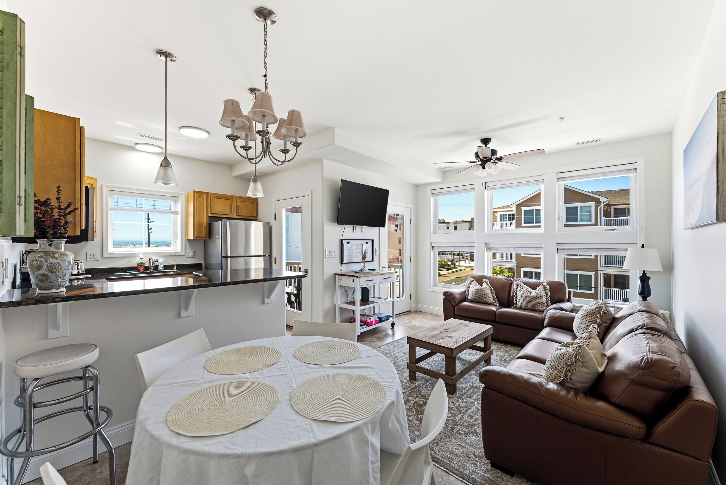 A modern kitchen with a dining area, a living room with a brown leather sofa, and a view of the neighborhood.