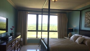 Ein Schlafzimmer mit einem großen Bett, einem Fernseher auf einer Holzkommode, einem Fenster mit Vorhängen und einer Lampe auf einem Nachttisch.