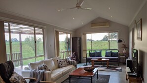 A spacious living room with a sofa, chairs, a coffee table, and a television. Large windows offer views of a green landscape.