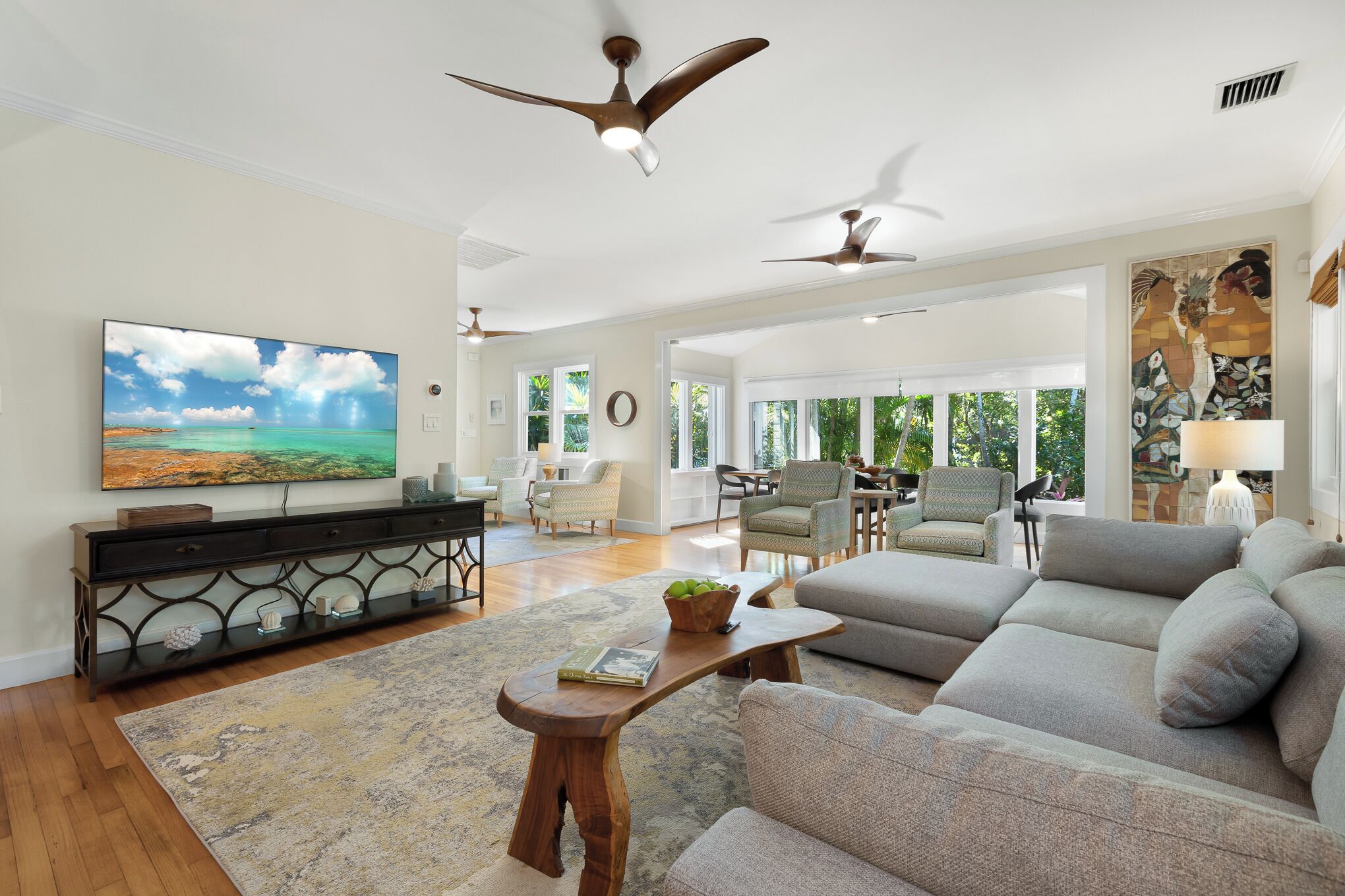 A spacious living room with a large flat-screen TV, a wooden coffee table, a grey sofa, and two ceiling fans.