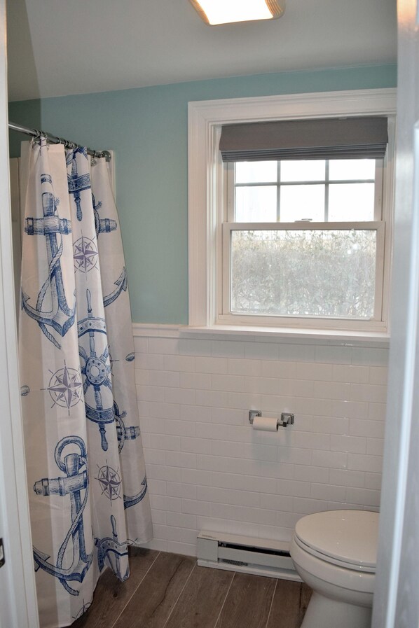A bathroom with a white tiled wall, a window with a view, a white toilet, and a shower curtain with nautical designs.