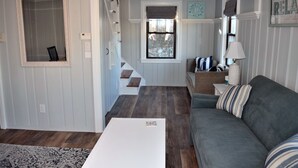 Una sala de estar con un sofá gris, una mesa de centro blanca y una alfombra estampada. Se ve una escalera que conduce a otra habitación, y una ventana con vistas a árboles.