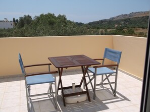 A balcony with a table and two chairs, a white cushion, and a view of a hilly landscape.