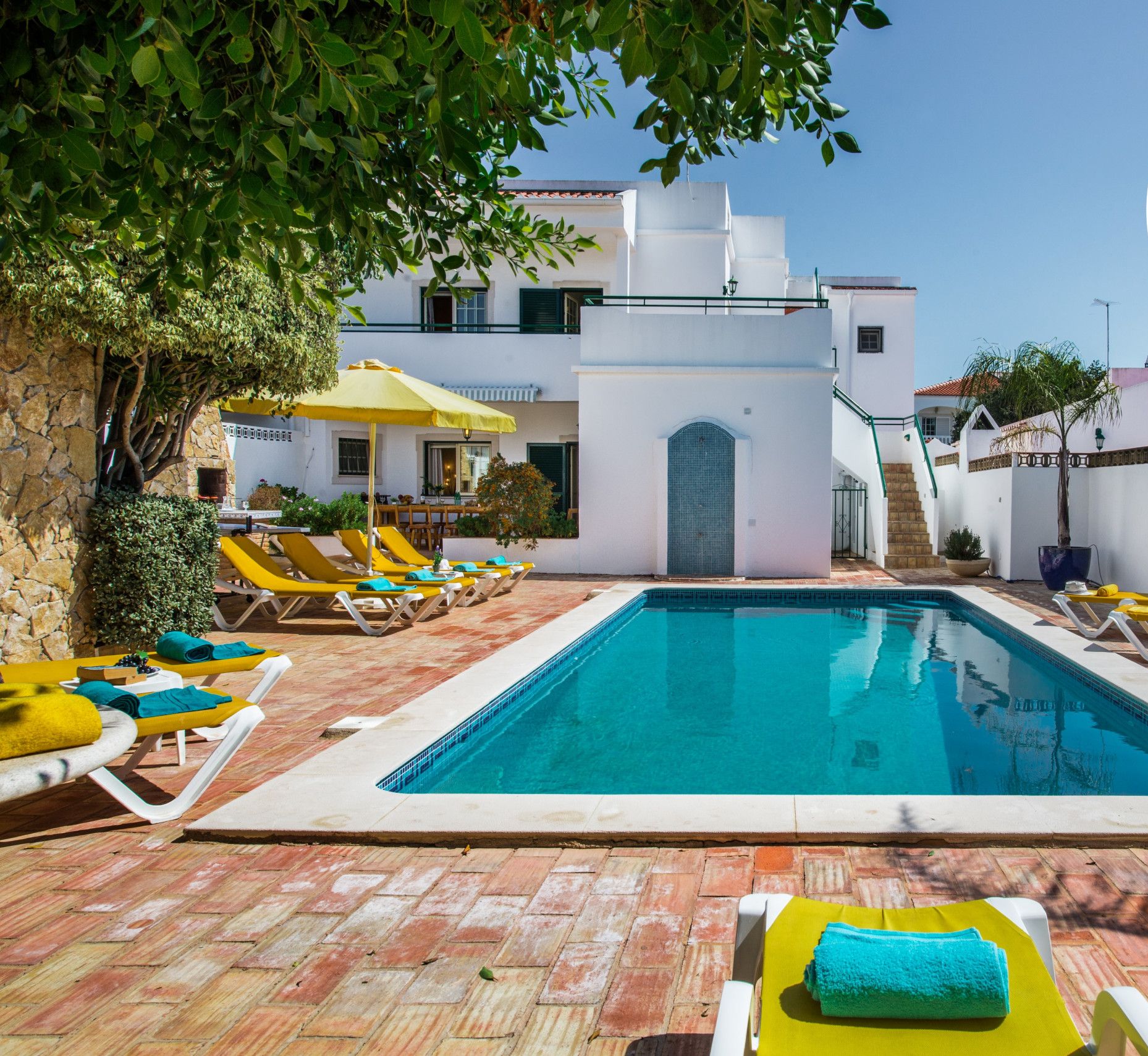 Une piscine entourée de chaises longues et de parasols, elle-même bordée d’un bâtiment blanc doté d’un toit en tuiles.