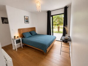 A bedroom with a wooden bed, a bedside table with a lamp, a small plant, and a view of greenery outside. - Bright wooden house 1 km from beaches and Aber Wrac'h (Landéda)
