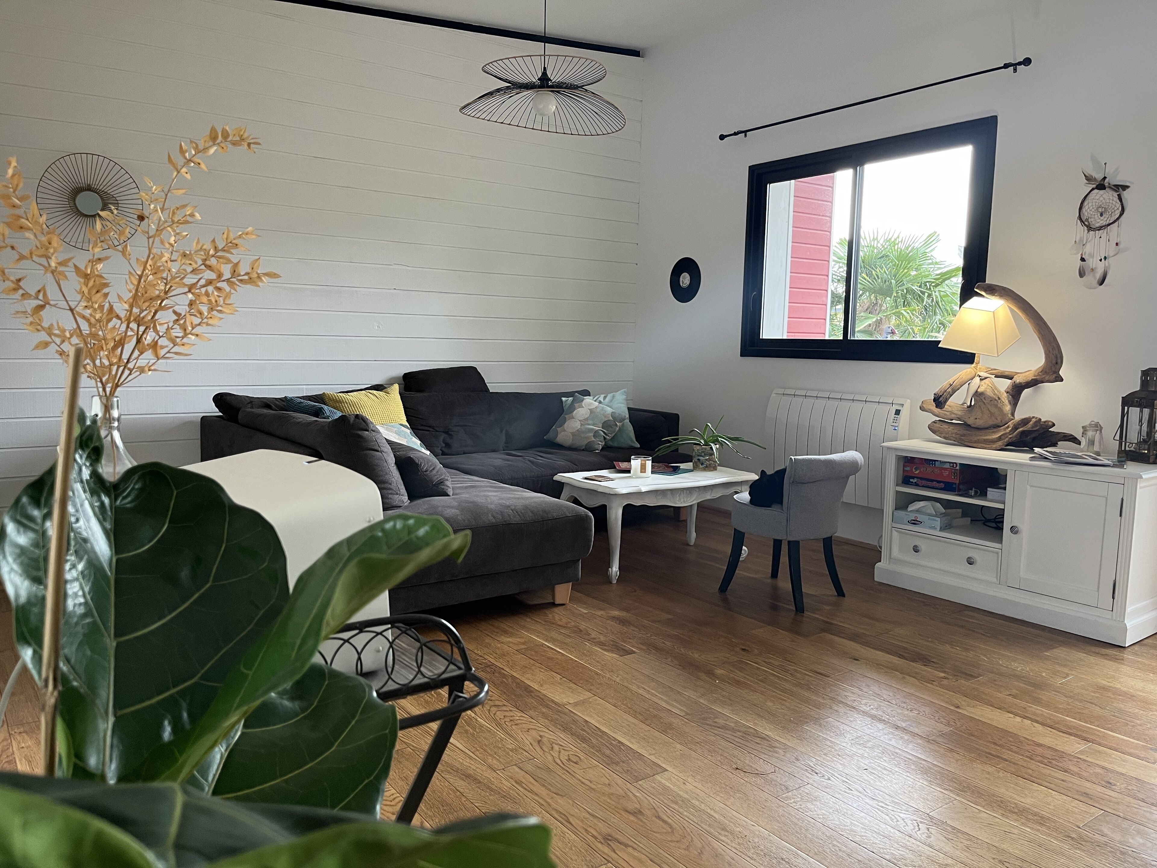 Una sala de estar moderna con un sofá seccional gris, una mesa de centro blanca y un sillón gris. Se aprecia una ventana grande con vistas a la vegetación, un armario blanco con una lámpara rústica y un ventilador de techo negro.
