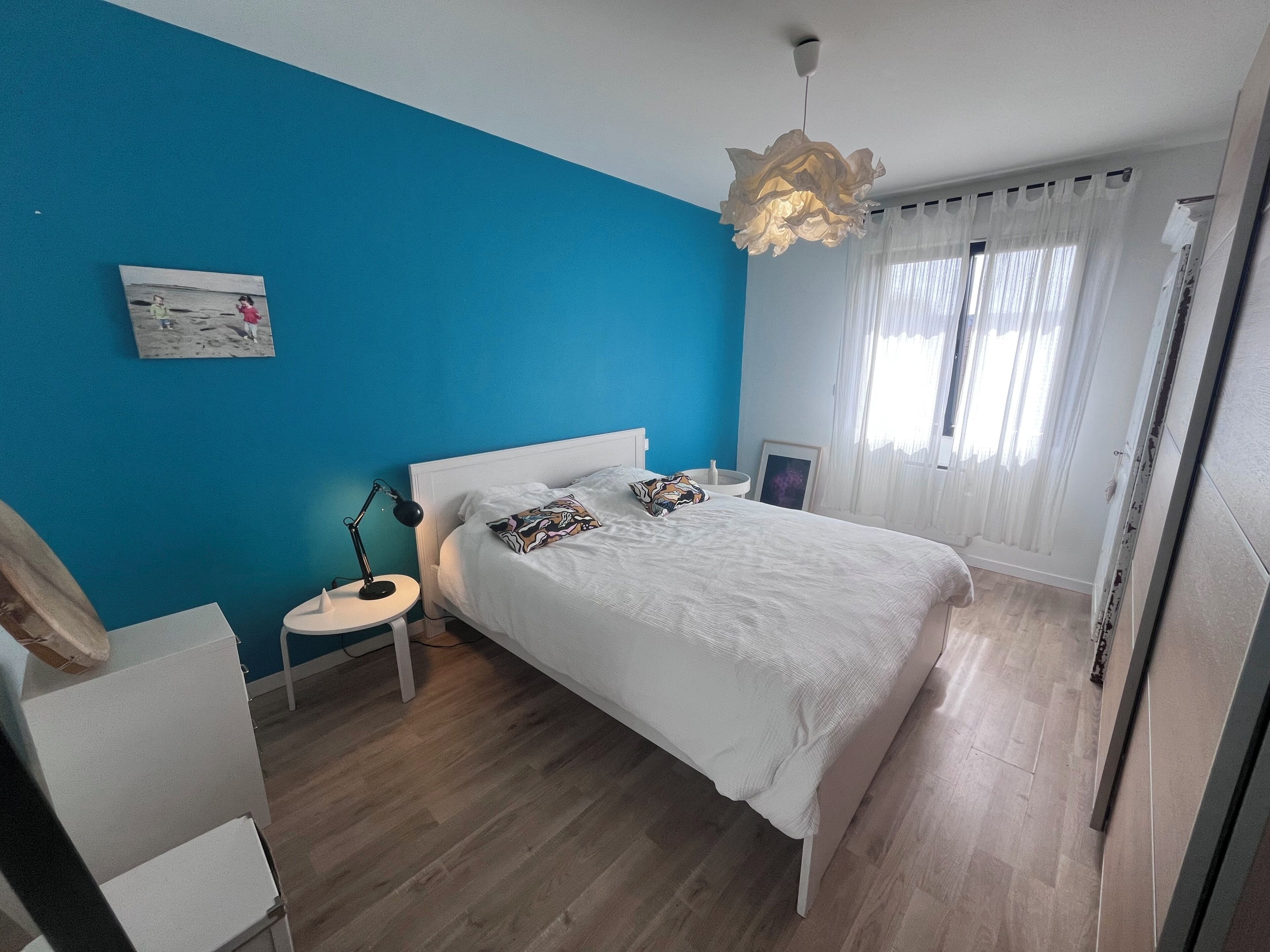 A bedroom with a bed, a nightstand, a lamp, a picture on the wall, and a window with curtains.