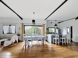 Et moderne køkken med en central ø, et spiseområde med bord og stole, og udsigt til grønne omgivelser.