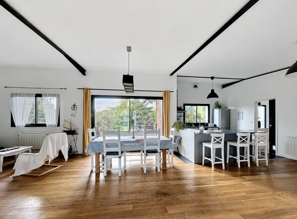 A modern kitchen with a central island, a dining area with a table and chairs, and a view of greenery outside.