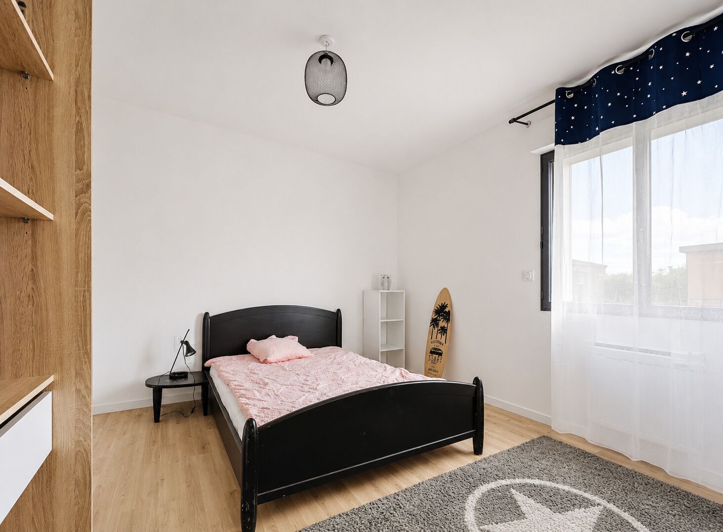 A bedroom with a bed, a nightstand, a lamp, a surfboard, a shelf, and a window with curtains.