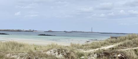 Eine Küstenlandschaft mit Sandstrand, grasbewachsenen Dünen und einem fernen Leuchtturm.