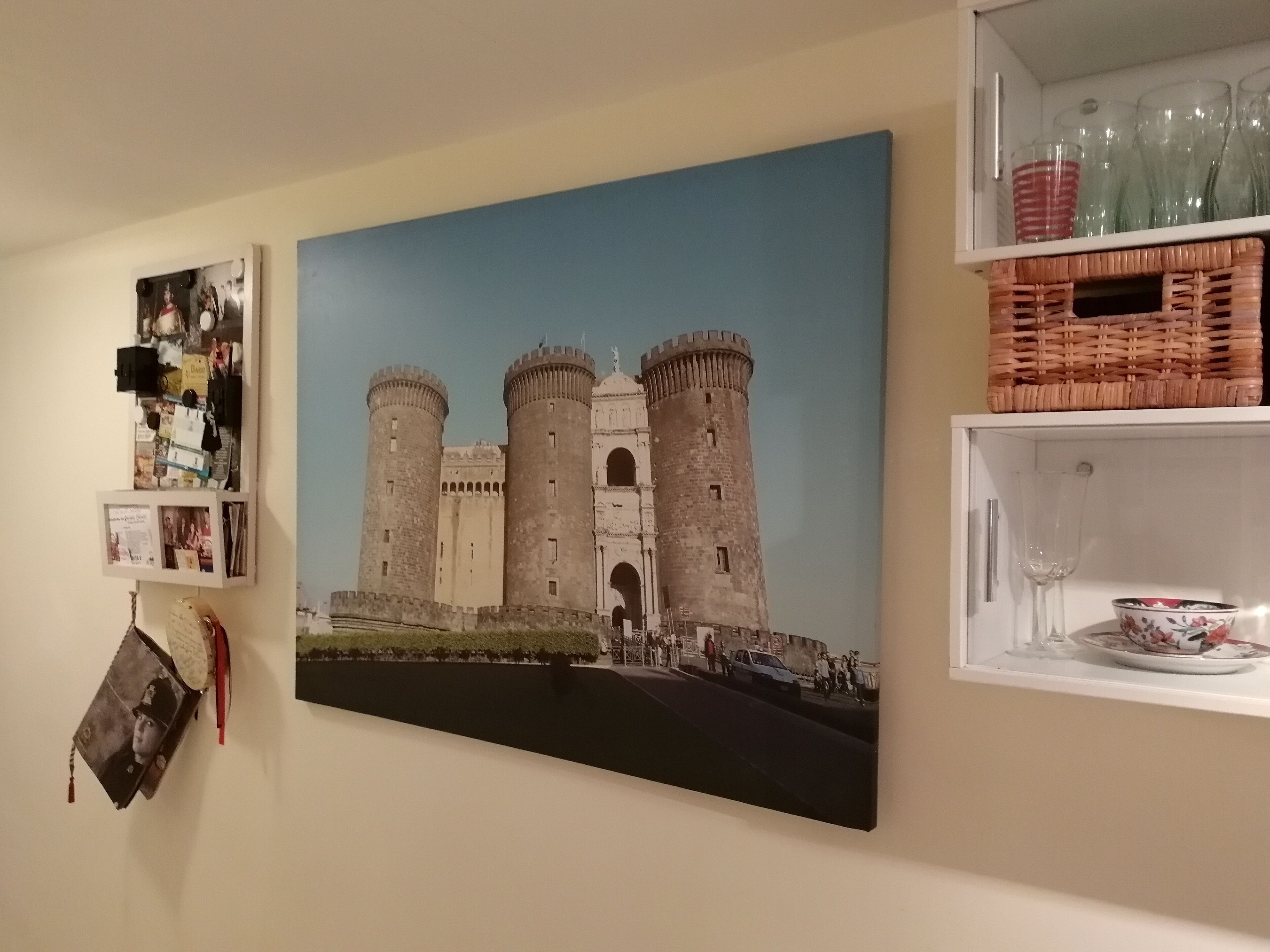 A wall with a large painting of a castle, a shelf with glassware, and a small framed photo.