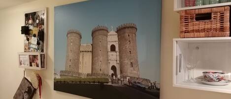 A wall with a large painting of a castle, a shelf with glassware, and a small framed photo.