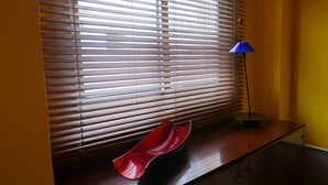 Een kamer met een bureau, een rode stoel, een lamp en een raam met zonwering.