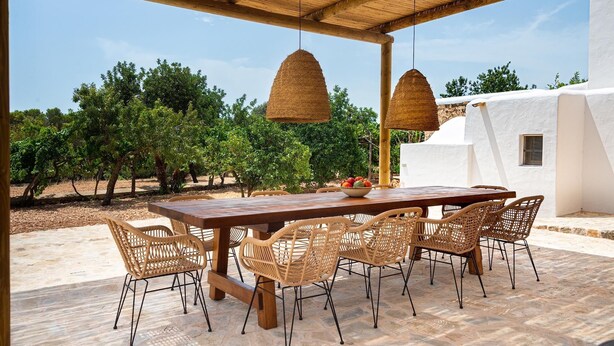 An outdoor dining area with a wooden table and wicker chairs, surrounded by a white building and trees.