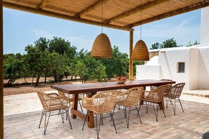 An outdoor dining area with a wooden table and wicker chairs, surrounded by a white building and trees. - House / Villa - Ibiza (Ibiza)