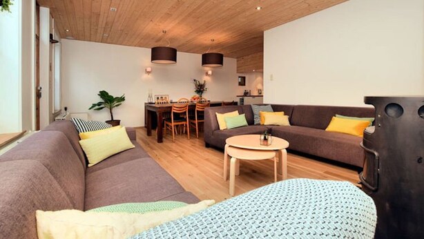 A modern living room with a wooden ceiling, a sofa set, a dining table, and a kitchen area in the background. - Vacation home Martha (Veurne)