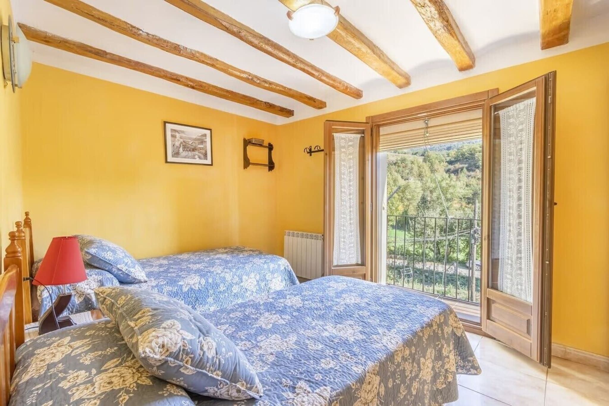 A bedroom with two beds, a red lamp, a framed picture on the wall, and a view of greenery through a sliding door.