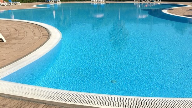 A clear blue swimming pool with lounge chairs and umbrellas, surrounded by greenery and hills.