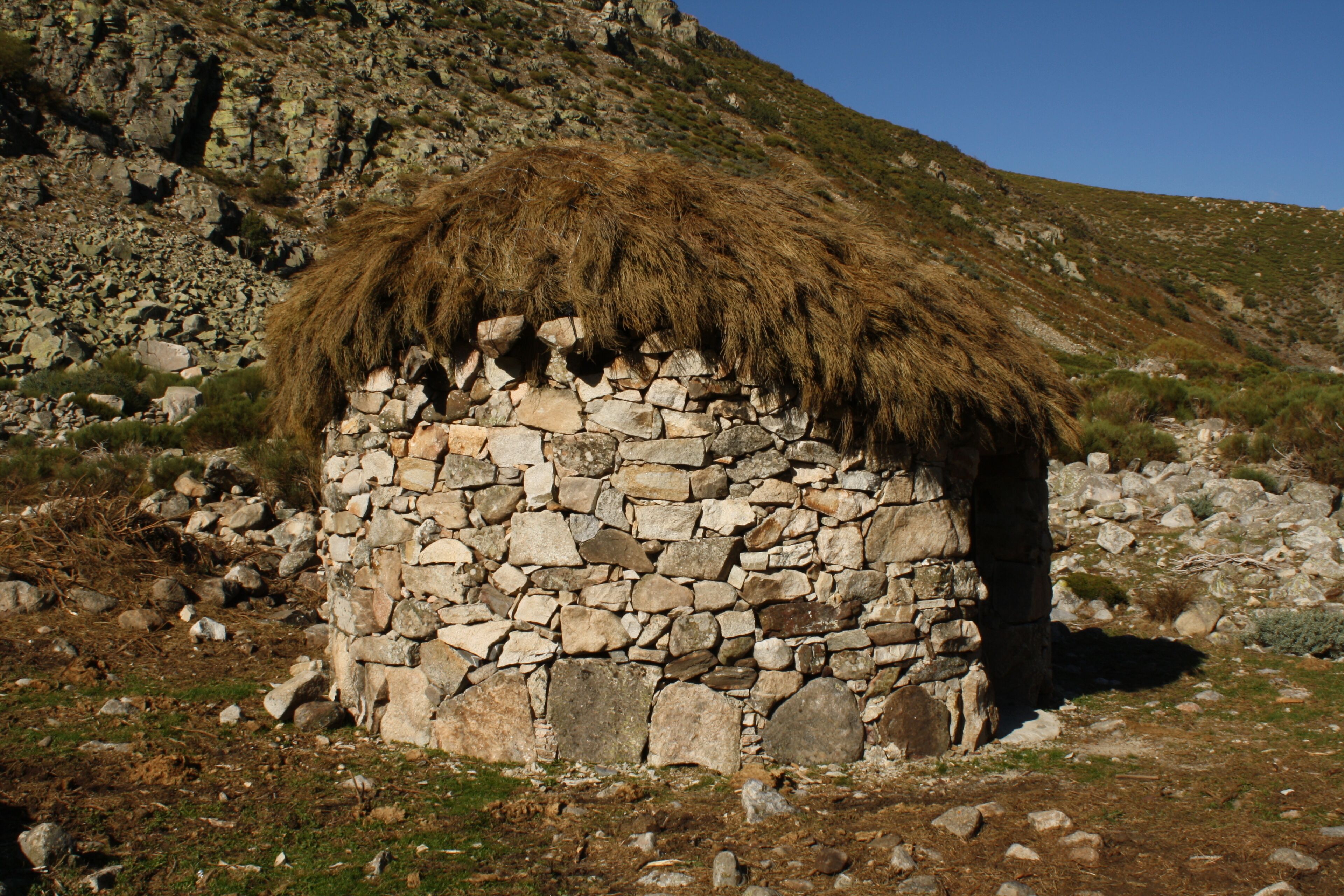 A stone hut with a thatched roof, surrounded by rocky terrain and sparse vegetation.