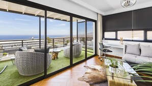 Una sala moderna con un amplio ventanal que da a un balcón con vista al océano. Hay dos sillones grandes de fibra natural, un tapete de cuero y un sofá blanco.