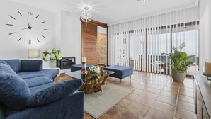 Una sala moderna con un sofá azul, una mesa de centro de madera, un reloj de pared grande y un balcón con vistas.