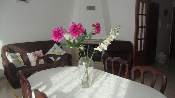 A dining table with a vase of flowers, surrounded by wooden chairs, in a room with a brown sofa and a fireplace.