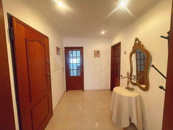 Un pasillo con una puerta de madera, una ventana, una mesa redonda con mantel blanco y un espejo en la pared.