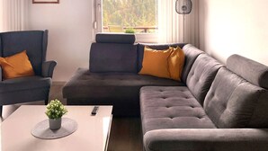 Un soggiorno moderno con un divano grigio, due poltrone, un tavolino da caffè con una pianta e vista su un lago attraverso una finestra.