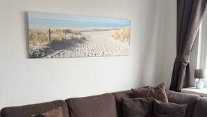 A living room with a brown sofa, a framed beach photograph, and a lamp on a side table. - Luxury vacation rental centrally located and close to the beach. Wi-Fi free. (Norderney)