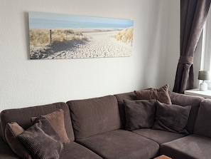 A living room with a brown sofa, a framed beach photograph, and a lamp on a side table.