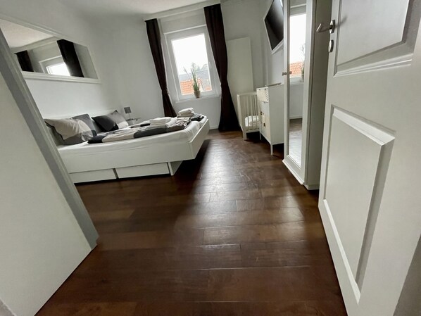 A narrow hallway leading to a bedroom with a bed, a radiator, and a window.